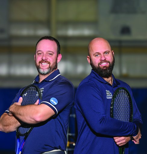 ODU tennis coaches Dominic Manilla and Dominik Mueller stand back-to-back on a tennis court. 