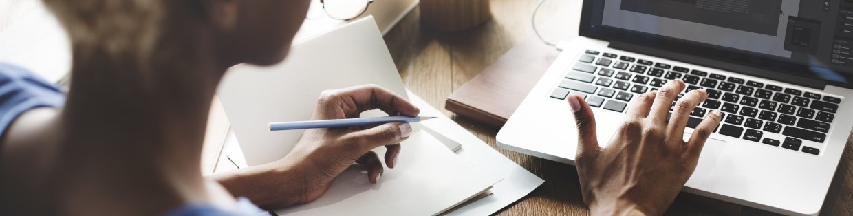 women typing on computer with a pen in her other hand on a pad of paper.