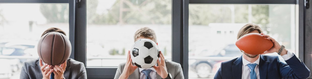 Three people sitting at chairs with objects in front of their face. One has a soccer ball, the other has a football, the last one holding a basketball. 