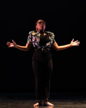 Ma’Kyia Frazier stands alone on stage, facing the audience with her arms open. 
