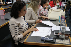 A woman sits at a desk in a classroom, reviewing documents and taking notes