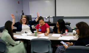  A group of students collaborate around a table in a classroom, with laptops,&nbsp;papers and a coffee cup on the surface.