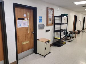 Hallway in a building. Photo is focused on one door that leads to the autoclave facility. The door is surrounding by a bulletin board, black shelves, a small cabinet, and many flyers on the walls.