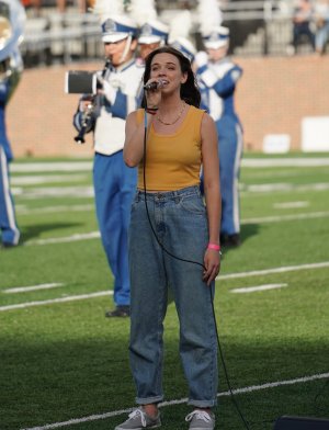 A woman sings into a microphone while standing on a football field. 