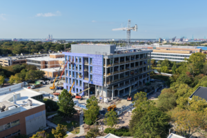 Aerial view of the new biological sciences building under construction on ODU campus, with cranes and construction equipment surrounding the site and the Norfolk waterfront and port visible in the distance.