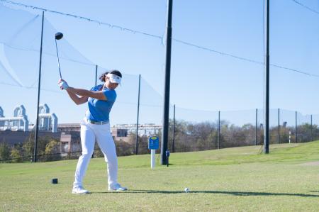 female student in a light blue polo, white pants, and white visor swinging her golf club on a golf course