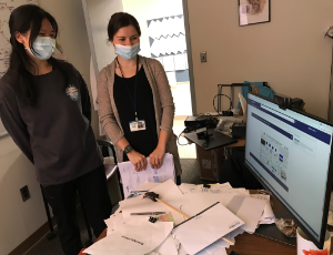 Two masked women standing next to a messy desk holding a com