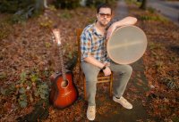 An image of Dennis Northerner seated on a wooden chair outside with a drum under one arm and a guitar standing upright beside him.