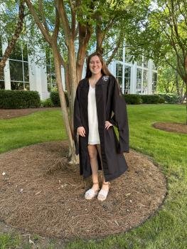 Bethany is standing in her graduation cap and gown underneath a tree.