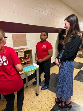 Student in red STEM Academy shirt presenting project to another student and a teacher.