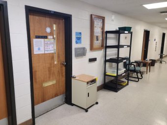 Hallway in a building. Photo is focused on one door that leads to the autoclave facility. The door is surrounding by a bulletin board, black shelves, a small cabinet, and many flyers on the walls.